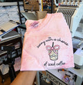 Pink tank top with text and graphic held in a workshop setting.but-first-coffee-shirt