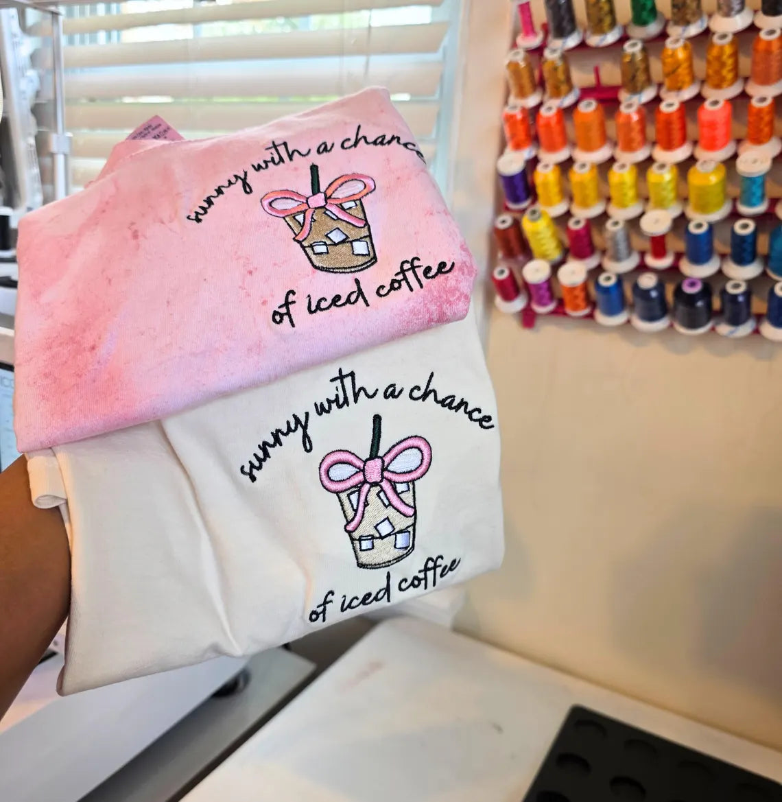 Two t-shirts with a design of iced coffee and text held in front of spools of .but-first-coffee-shirtthread.