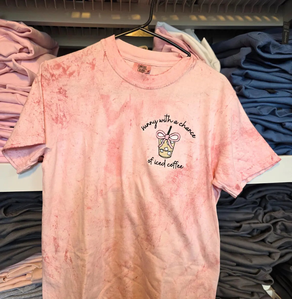 Pink tie-dye t-shirt with text and graphic on a hanger among folded shirts.but-first-coffee-shirt