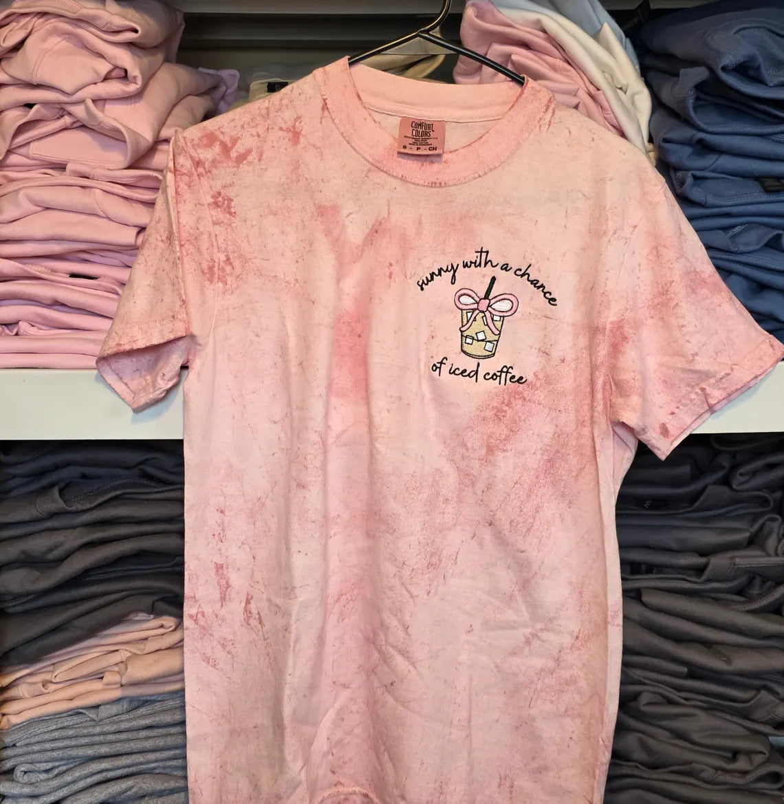 Pink tie-dye t-shirt with text and graphic on a hanger surrounded by folded -but-first-coffee-shirtclothes.