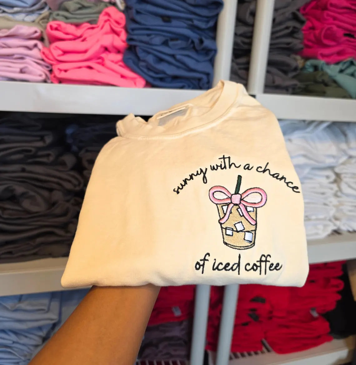 Beige t-shirt with 'sunny with a chance of iced coffee' text and graphic held in front of a display of colorful t-shirts.but-first-coffee-shirt