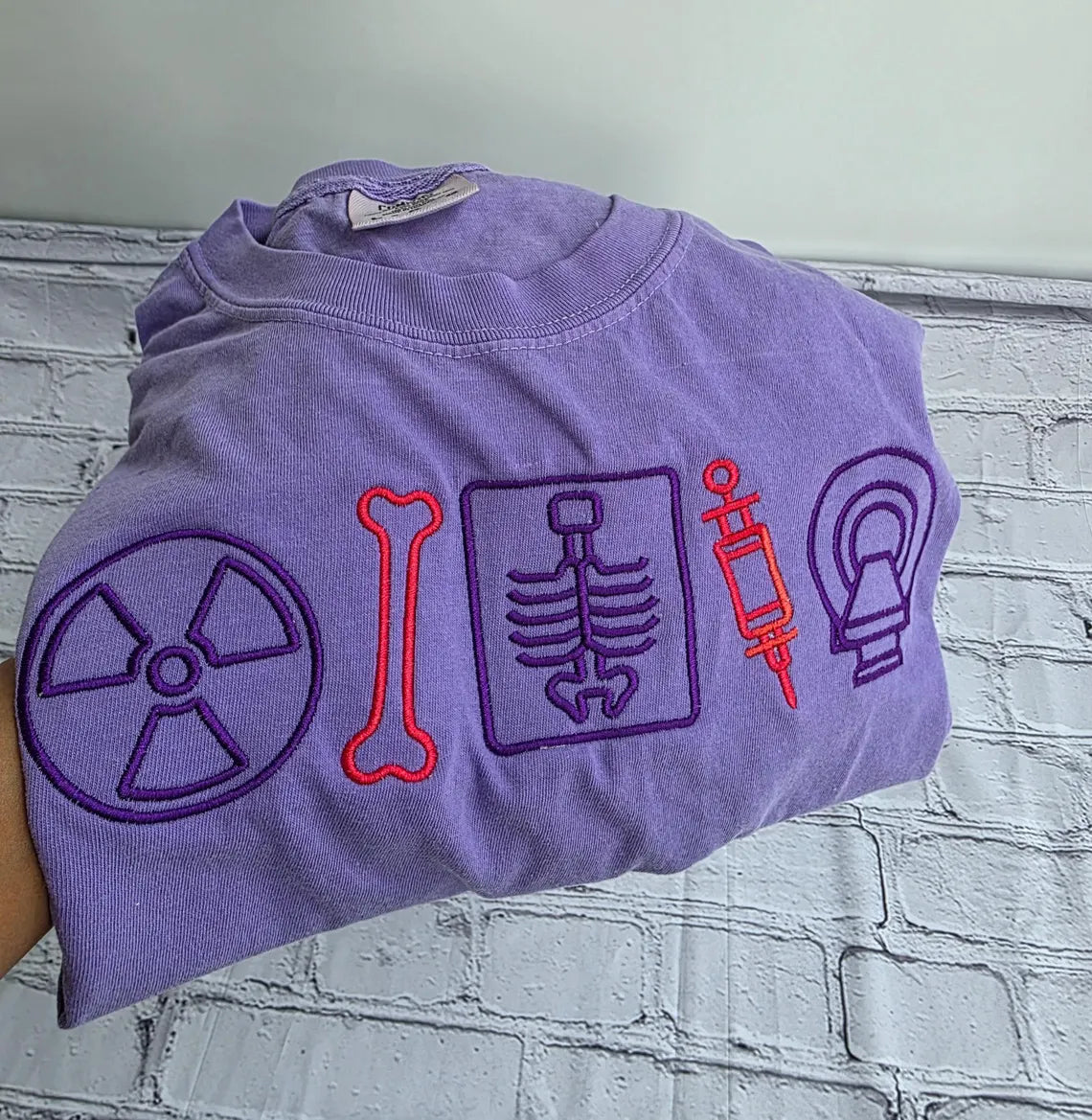 Folded purple t-shirt with embroidered symbols on a textured surface.radiologist-t-shirt