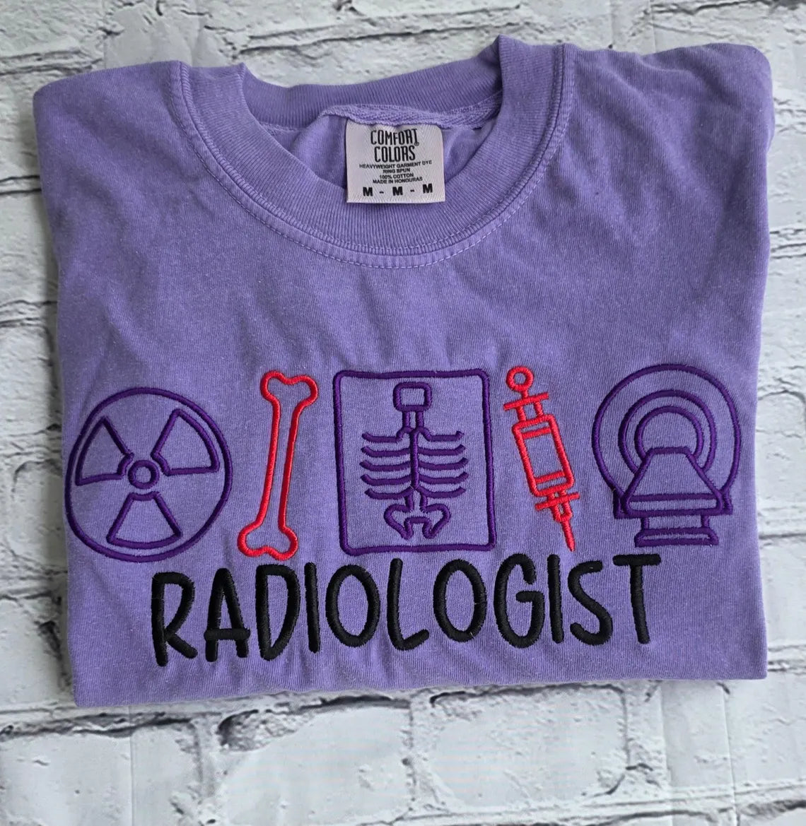 Folded purple t-shirt with radiology-themed graphics and the word 'Radiologist' on a textured gray background.radiologist-t-shirt