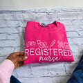 Pink sweatshirt with 'Registered Nurse' text and medical symbols on a textured surface.nurse-embroidered-sweatshirt