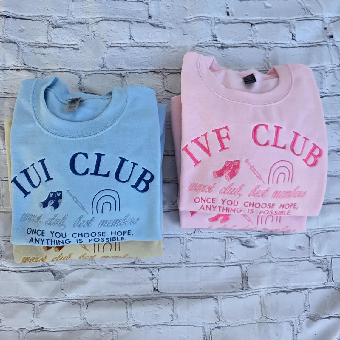Two folded sweatshirts, one blue and one pink, with text on a brick wall background. ivf-sweatshirt