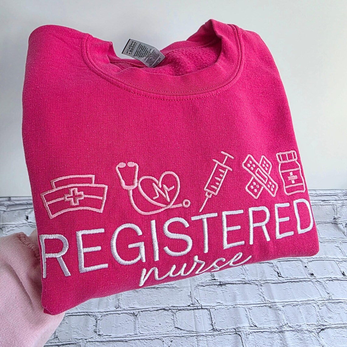 Pink sweatshirt with 'Registered Nurse' text and icons on a white background.nurse-embroidered-sweatshirt