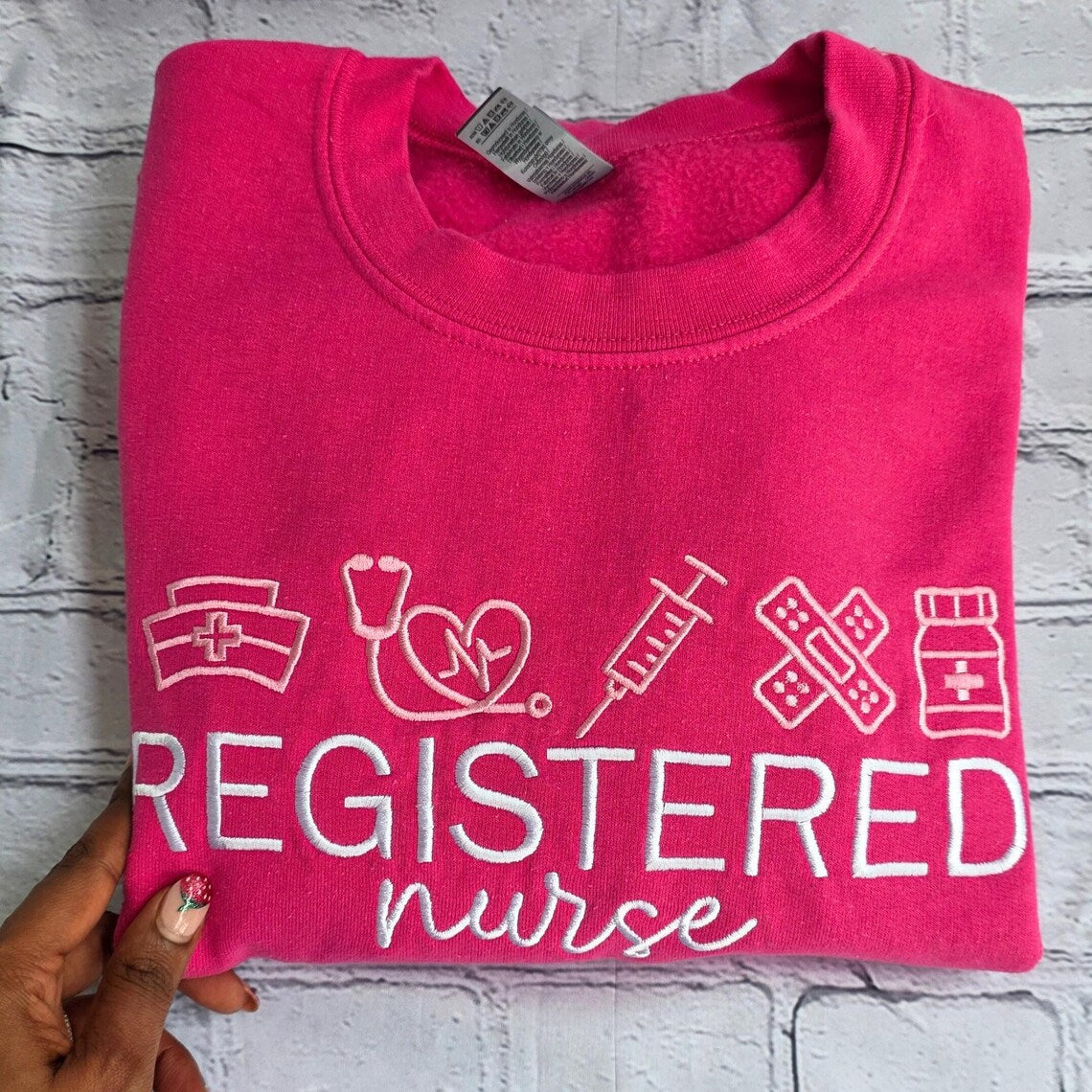 Folded pink sweatshirt with 'Registered Nurse' text and symbols on a textured surface.nurse-embroidered-sweatshirt