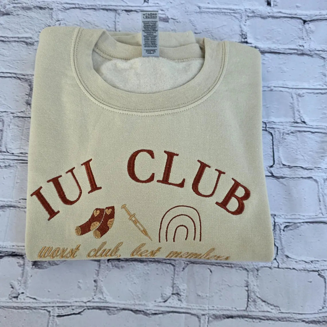 Folded beige sweatshirt with 'IUI Club' text and graphics on a textured white background. ivf-sweatshirt