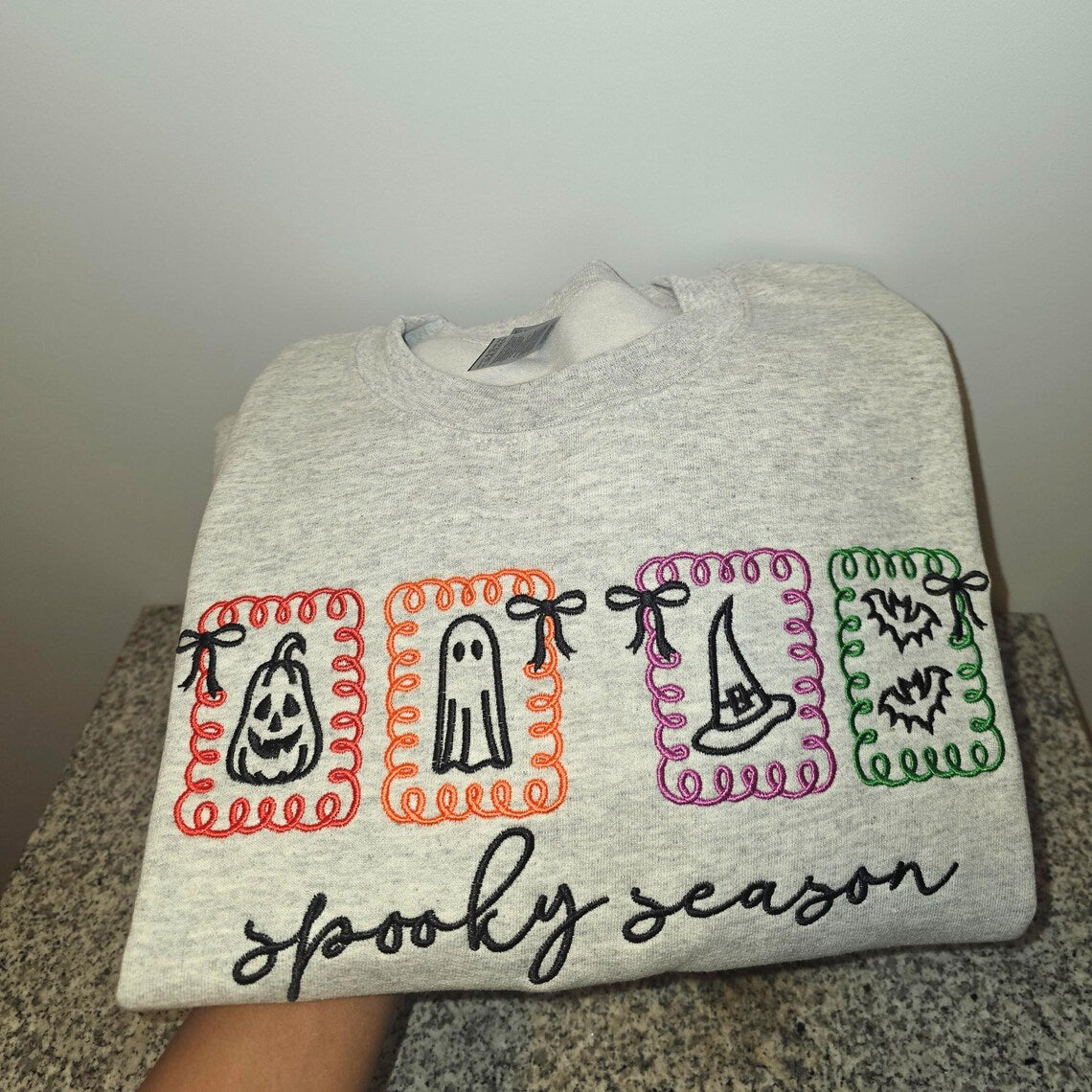 Gray sweatshirt with Halloween-themed designs and 'spooky season' text on a neutral background.halloween-embroidery-sweatshirt