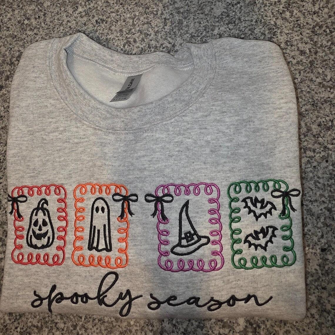 Folded gray sweatshirt with Halloween-themed embroidery on a stone surface.halloween-embroidery-sweatshirt