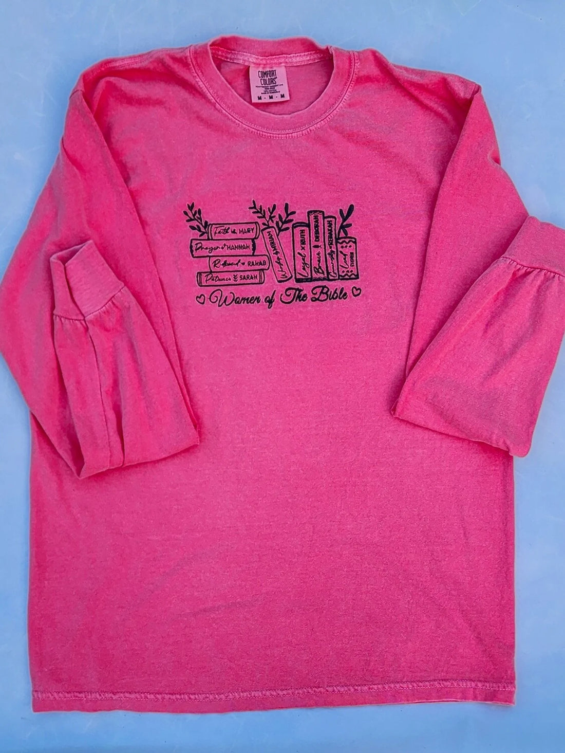 A woman wearing a pink long-sleeved t-shirt with embroidered text that reads 'Women of the Bible' and includes images of various women mentioned in the Bible.women-of-the-bible-shirt