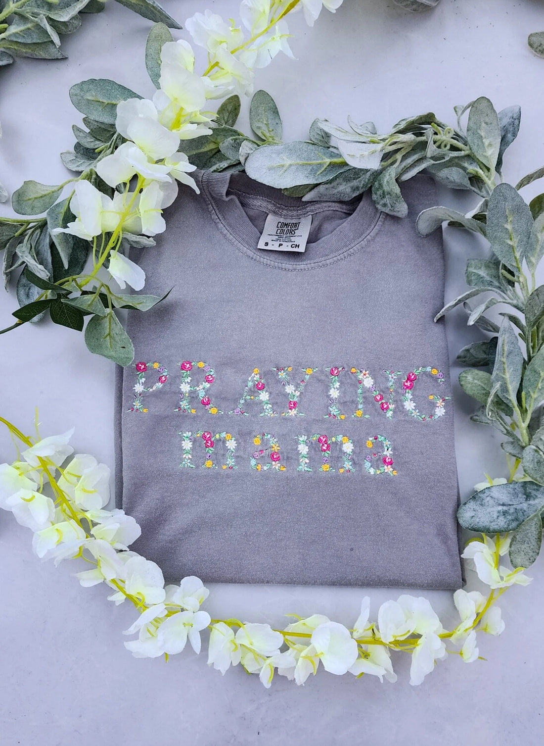 A long sleeve gray t-shirt with the text 'PRAYING MAMA' embroidered in colorful letters, displayed flat with a floral arrangement around it.praying-mama-shirt