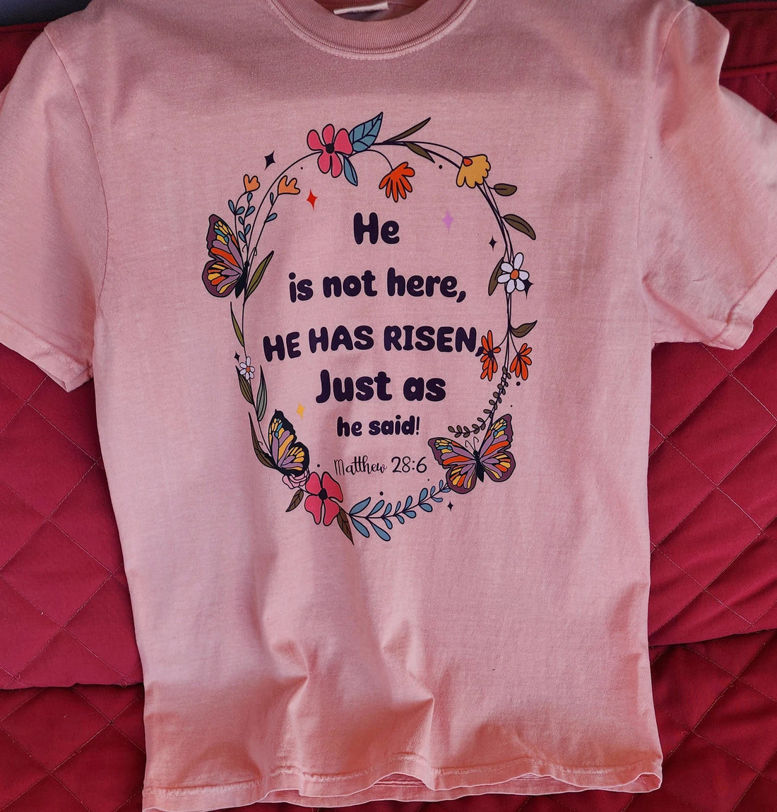 Stacked pink t-shirts with the text 'He is not here, HE HAS Risen' printed on them, placed on a red surface.