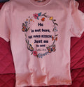 Stacked pink t-shirts with the text 'He is not here, HE HAS Risen' printed on them, placed on a red surface.