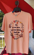Stacked pink t-shirts with the text 'He is not here, HE HAS Risen' printed on them, placed on a red surface.