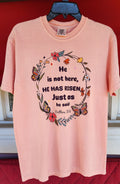Stacked pink t-shirts with the text 'He is not here, HE HAS Risen' printed on them, placed on a red surface.