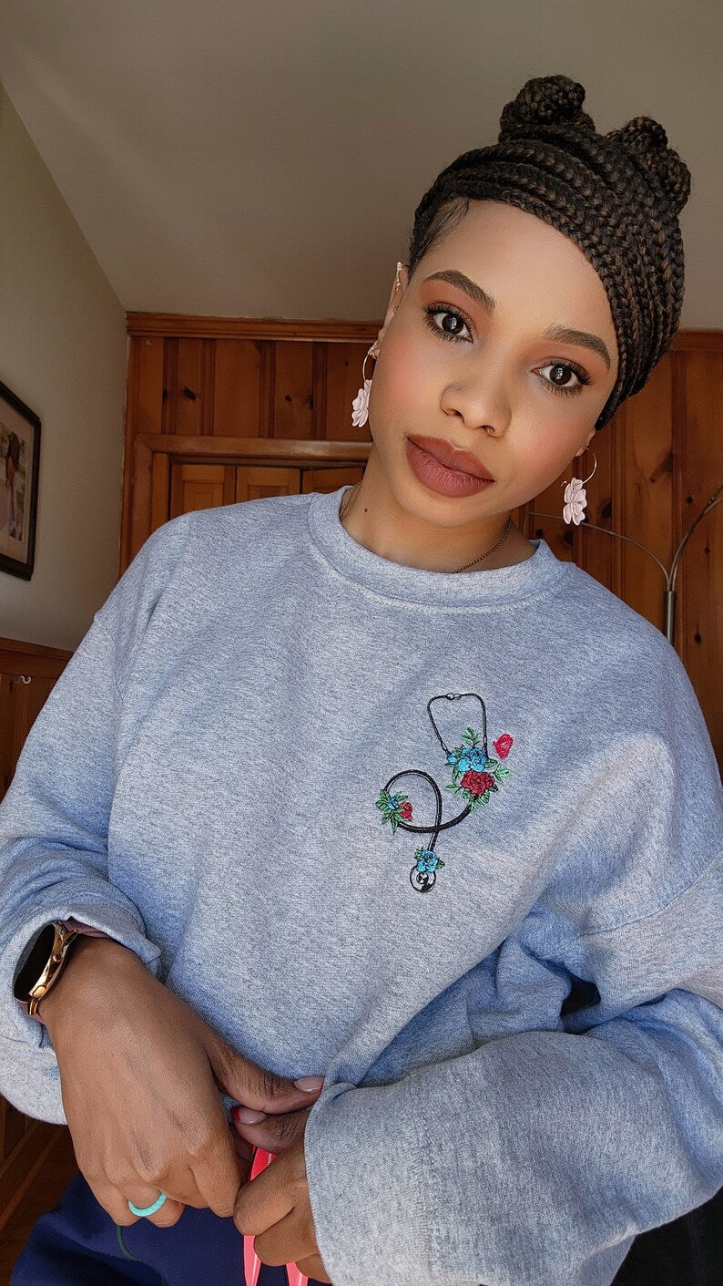 A person wearing a gray sweatshirt with an embroidered stethoscope and floral pattern on the chest.