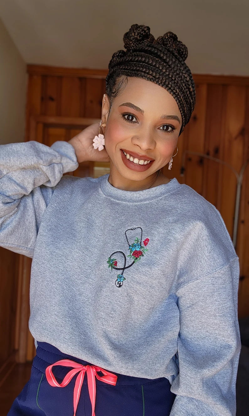 A person wearing a gray sweatshirt with an embroidered stethoscope and floral pattern on the chest.