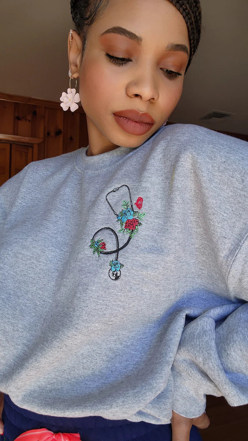 A person wearing a gray sweatshirt with an embroidered stethoscope and floral pattern on the chest.