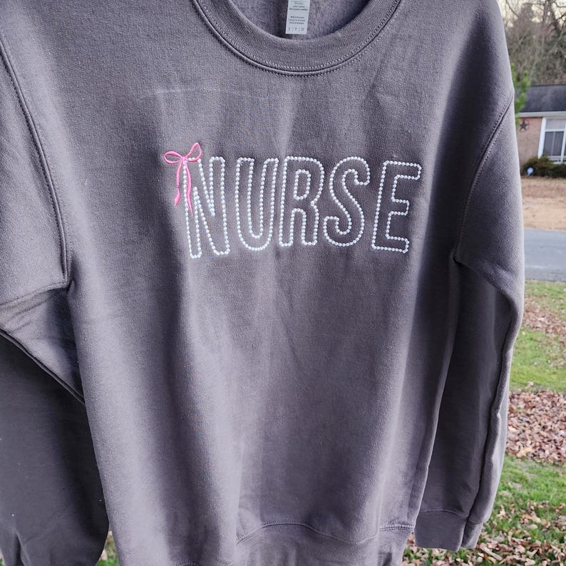 A collection of custom embroidered grey nurse shirts with various titles and names embroidered on the left side of the chest.