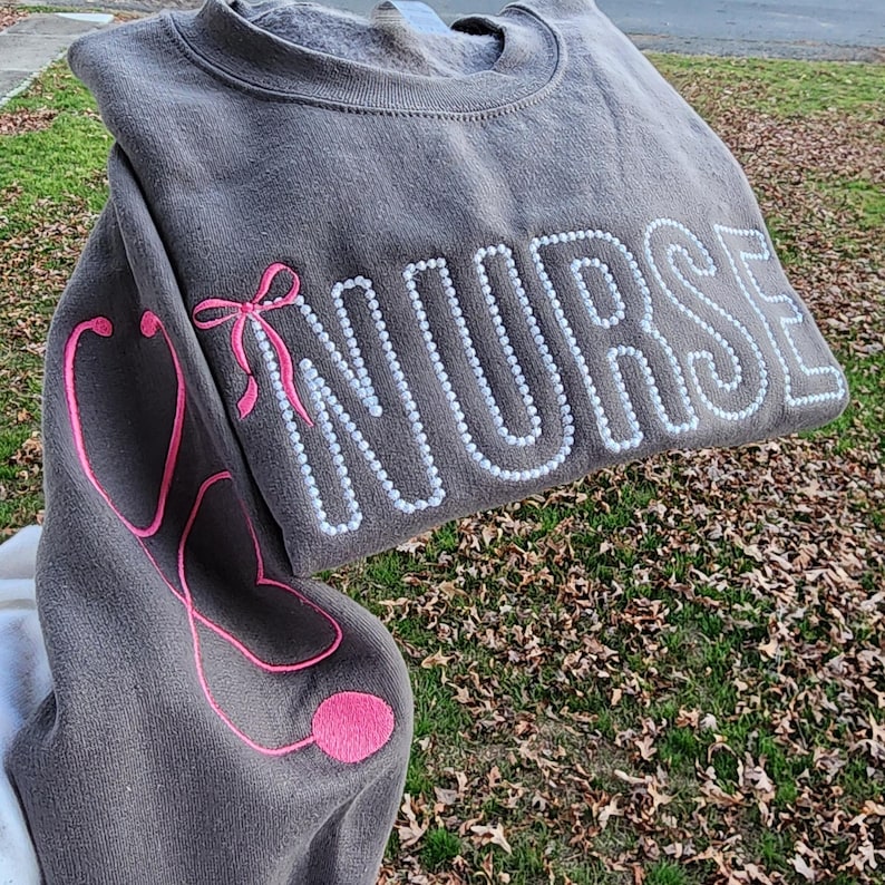 A collection of custom embroidered grey nurse shirts with various titles and names embroidered on the left side of the chest.
