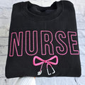 Black sweatshirt with embroidered pink text spelling 'NURSE' and a heart-shaped bow design underneath.