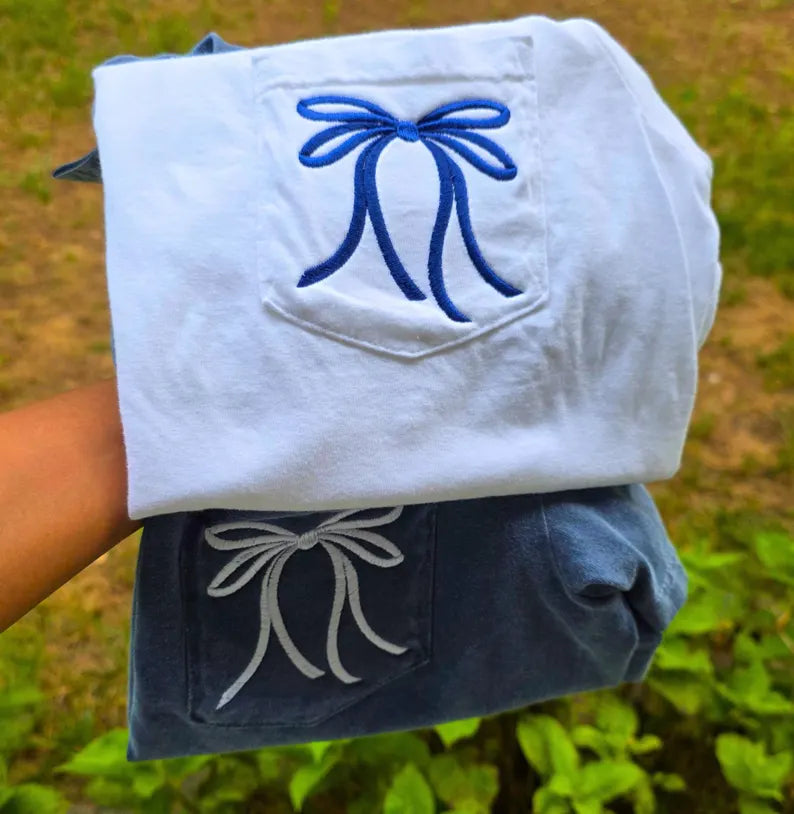 White shirt with blue bow design held by a person against a natural background