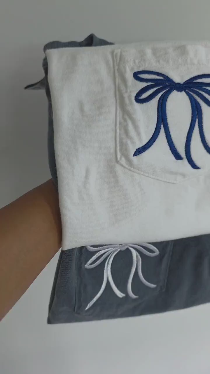 White shirt with blue embroidered bow and navy pants with white embroidered bow on a plain background